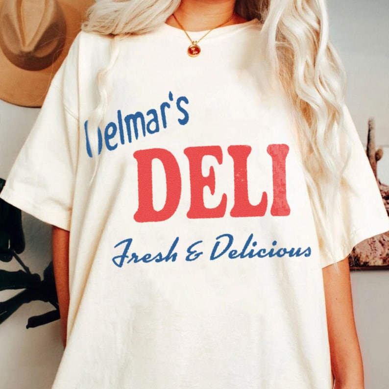 May include: An off-white short-sleeve t-shirt featuring the blue text Delmar's, the large red text DELI, and the blue text Fresh & Delicious. The shirt is worn with a gold chain necklace featuring a small round pendant.