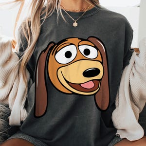 Pu&ograve; includere: Felpa grigio scuro con un'illustrazione a cartoni animati di un cane sorridente. Il cane ha orecchie marroni, faccia beige e lingua rosa. La felpa &egrave; in stile casual.
