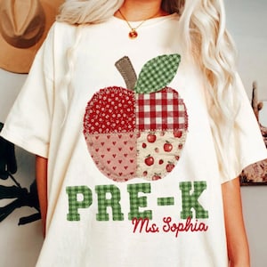 May include: A cream-colored t-shirt featuring a large apple graphic composed of four distinct fabric-style patterns: red floral, red and white gingham, pink with small hearts, and cream with tiny red apples. The text "PRE-K" and "Ms. Sophia" appear below.