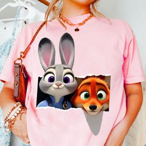 May include: Pink t-shirt featuring a graphic of Judy Hopps and Nick Wilde from Zootopia. The characters are depicted in a torn paper effect. The shirt is a solid pink color and has short sleeves.