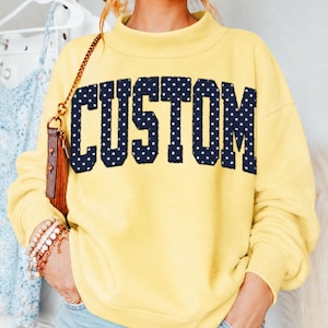 May include: A pale yellow long-sleeve sweatshirt featuring a mock neck collar. The word CUSTOM is printed across the chest in large, dark navy blue block letters with a white polka dot pattern. The sweatshirt has long sleeves with ribbed cuffs.