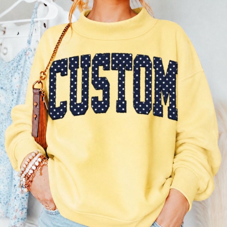 May include: A pale yellow long-sleeve sweatshirt featuring a mock neck collar. The word CUSTOM is printed across the chest in large, dark navy blue block letters with a white polka dot pattern. The sweatshirt has long sleeves with ribbed cuffs.