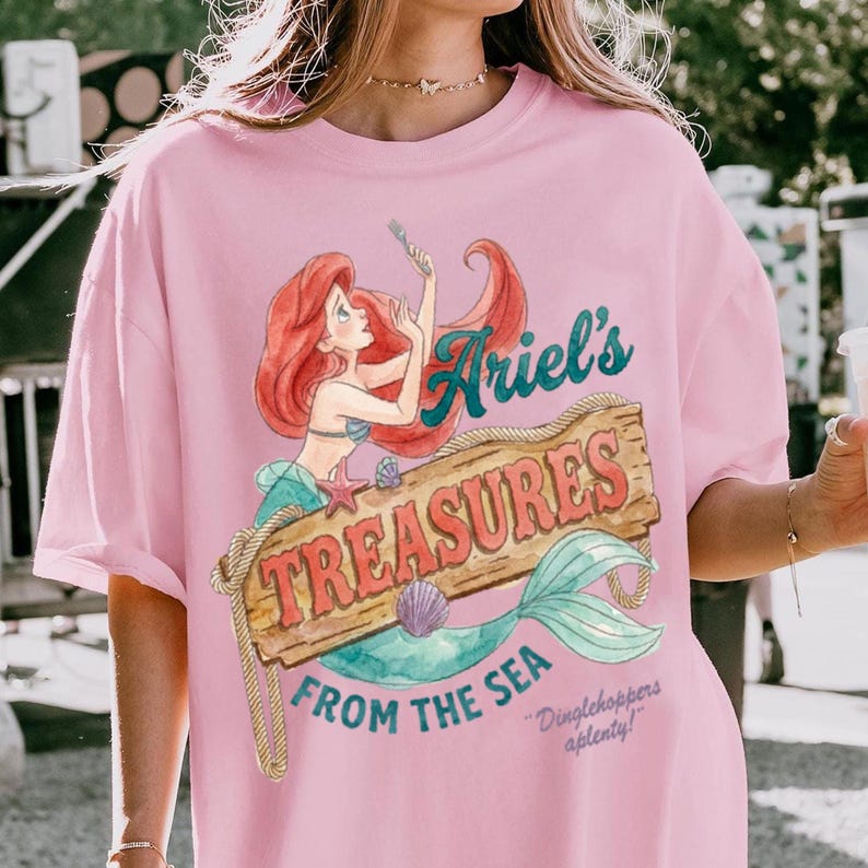 May include: Pink t-shirt featuring a watercolor illustration of Ariel from The Little Mermaid. The design includes the text "Ariel's Treasures From The Sea" on a wooden sign, with a mermaid and seashell. The shirt also has the text "Dinglehoppers aplenty!"
