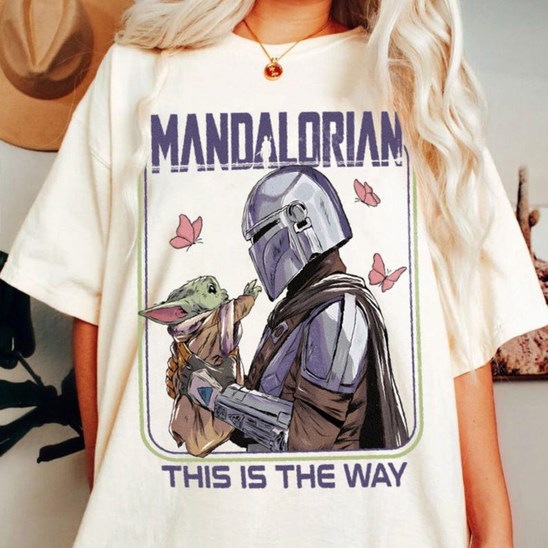 May include: Cream-colored t-shirt featuring a graphic of The Mandalorian and Grogu, with the text "MANDALORIAN" above the image and "THIS IS THE WAY" below. The design includes pink butterflies.
