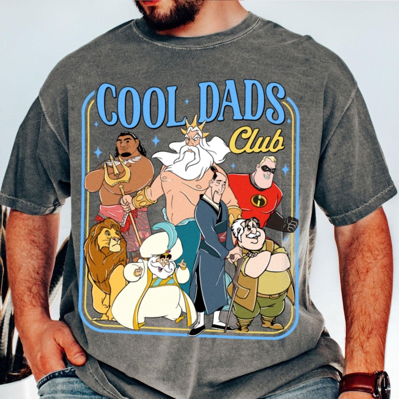 May include: Grey t-shirt with a graphic featuring cartoon characters and the text "COOL DADS CLUB" in blue and yellow. The design includes illustrations of various animated figures, set within a blue and yellow frame.