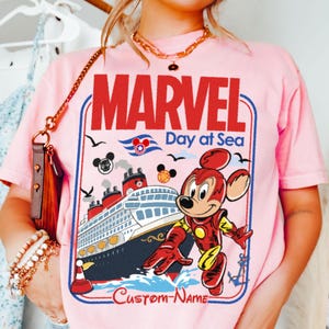 May include: Pink t-shirt featuring a graphic of a cruise ship with the words "MARVEL Day at Sea". The design includes a cartoon character in a superhero costume, Mickey Mouse ears, and the text "Custom Name".