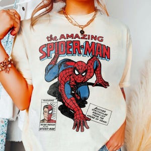 以下が含まれることがあります： ヴィンテージコミックのグラフィックが特徴のクリーム色のTシャツ。デザインには、赤と青で「the AMAZING SPIDER-MAN」というテキスト、追加のコミックテキスト、ピーター・パーカーの肖像画が含まれています。