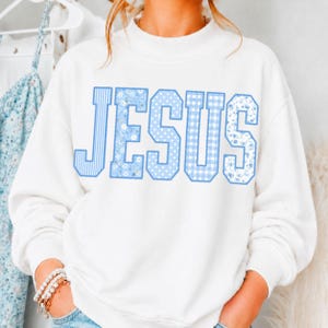 May include: A white long-sleeve sweatshirt featuring the word JESUS printed across the chest in large block letters. Each letter displays a different light blue pattern, including vertical stripes, small white flowers, polka dots, and gingham checks.