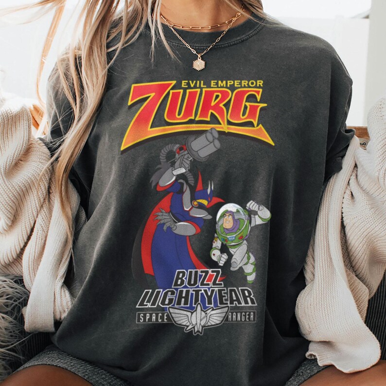 May include: Dark gray sweatshirt featuring a graphic of Evil Emperor Zurg and Buzz Lightyear from Toy Story. The design includes the text "EVIL EMPEROR ZURG" and "BUZZ LIGHTYEAR SPACE RANGER". The sweatshirt has a vintage, worn look.