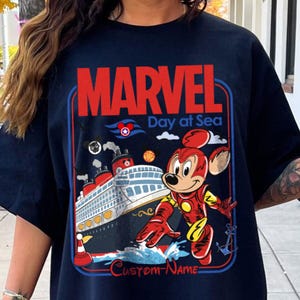 May include: Navy blue t-shirt featuring a graphic of Mickey Mouse in an Iron Man suit, a cruise ship, and the text "MARVEL Day at Sea". The design includes a red, white, and blue color scheme with the words "Custom-Name" at the bottom.
