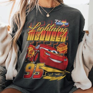 May include: Dark gray sweatshirt featuring a graphic of Lightning McQueen from the movie Cars. The design includes the text "Lightning McQueen" in yellow and red, the number "95", and the "Rust-eze" logo. The sweatshirt has a vintage, worn look.