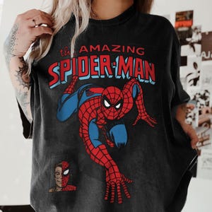以下が含まれることがあります： ダイナミックなポーズの、スパイダーマンの大きなグラフィックが特徴の黒いTシャツ。「the AMAZING SPIDER-MAN」の文字が赤と青でデザインされています。スパイダーマンの顔の小さな画像が左下にあります。