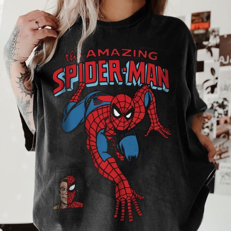 以下が含まれることがあります： ダイナミックなポーズの、スパイダーマンの大きなグラフィックが特徴の黒いTシャツ。「the AMAZING SPIDER-MAN」の文字が赤と青でデザインされています。スパイダーマンの顔の小さな画像が左下にあります。
