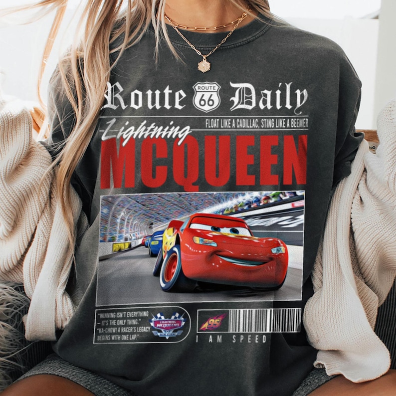 Op de afbeelding: Een grijs T-shirt met een grafische print van een rode raceauto met de tekst "Route Daily Lightning McQueen" en "Float like a Cadillac, Sting like a Beeemer". De grafiek bevat ook de tekst "I am Speed" en "Winning isn't everything - It's the only thing. Ka-Chow! A racer's legacy begins with one lap."