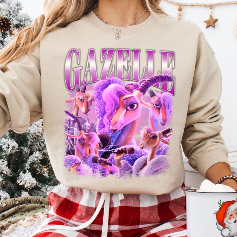 May include: Beige sweatshirt featuring a graphic of a purple gazelle with the word "GAZELLE" in a purple and white font. The gazelle has purple hair and is holding a microphone. The sweatshirt is paired with red and white plaid pajama pants.