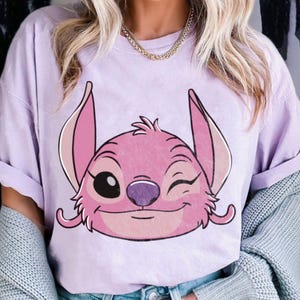 May include: A light purple t-shirt featuring a pink cartoon character with large ears and a winking expression. The character has a black eye, a purple nose, and a smiling mouth. The shirt has rolled-up sleeves.