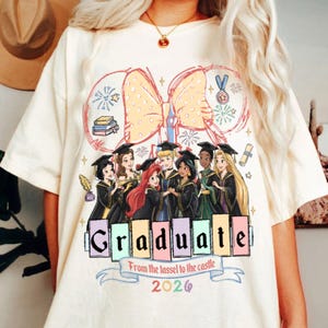 Puede incluir: Camiseta color crema con un gr&aacute;fico de princesas de Disney con atuendo de graduaci&oacute;n. El dise&ntilde;o incluye un gran lazo, fuegos artificiales, libros y la palabra "Graduate" con "From the tassel to the castle 2026" debajo.