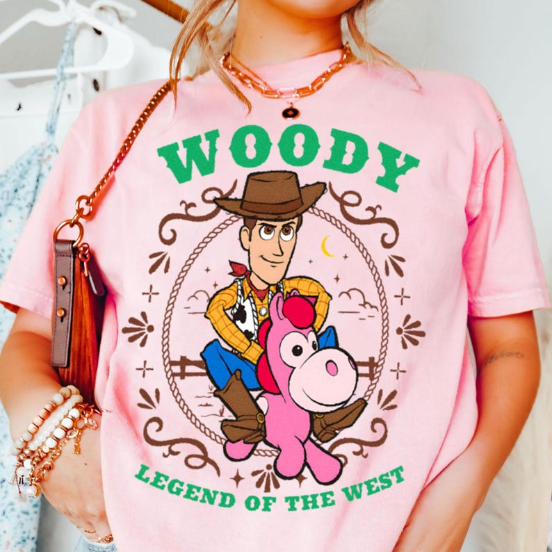 May include: A light pink t-shirt featuring a graphic of the character Woody riding a pink horse. The design includes a brown rope border, green text that reads WOODY and LEGEND OF THE WEST, and a small yellow crescent moon in the background.