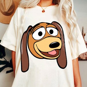Pu&ograve; includere: T-shirt color crema con un'immagine a cartone animato di Slinky Dog di Toy Story. La grafica mostra la testa di Slinky con orecchie marroni, muso beige e lingua rosa. La maglietta ha maniche corte e una vestibilit&agrave; ampia.