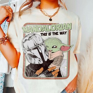 Puede incluir: Camiseta color crema con un gr&aacute;fico de The Mandalorian y Grogu, con el texto "MANDALORIAN THIS IS THE WAY" en verde y blanco. El dise&ntilde;o incluye una figura con casco y un personaje alien&iacute;gena verde.