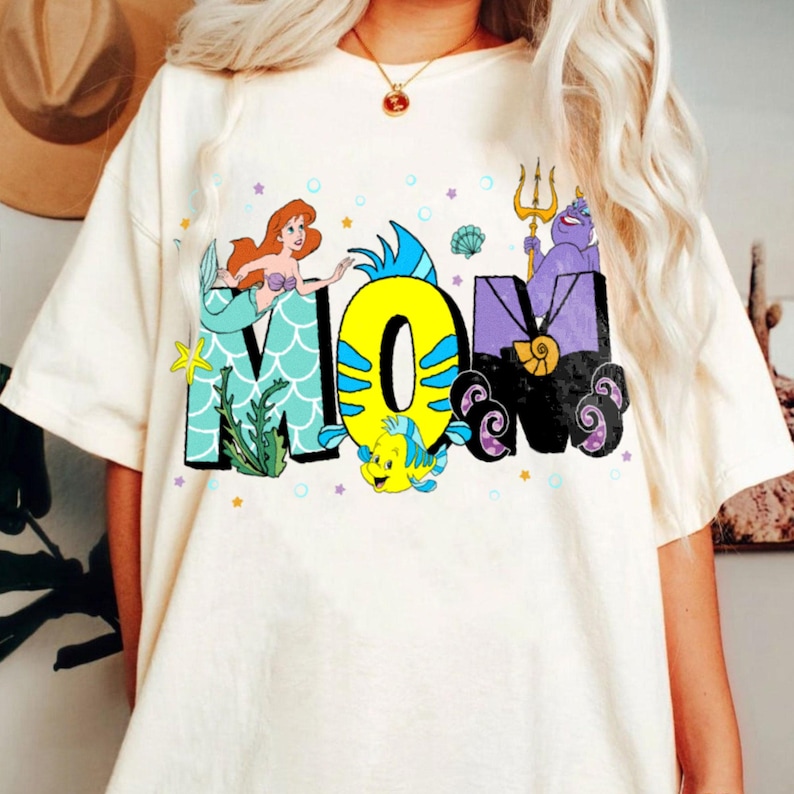 May include: White t-shirt with a colorful graphic of the word "MOM" featuring characters from the Little Mermaid. The graphic includes Ariel, Flounder, Ursula, and a seashell.
