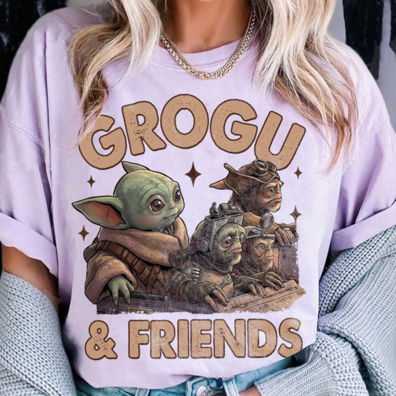 K&ouml;nnte beinhalten: Helllila T-Shirt mit dem Text "GROGU & FRIENDS" in Braun. Die Grafik zeigt Grogu und andere Charaktere aus Star Wars. Das Design beinhaltet Sterne und einen Vintage-Look.