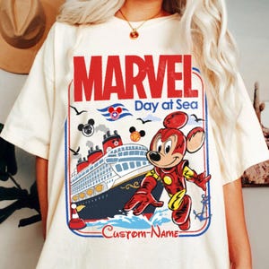 May include: Cream-colored t-shirt featuring a Marvel-themed design. The graphic includes the word "MARVEL" in red, a cruise ship, and a Mickey Mouse character in an Iron Man suit. The text "Day at Sea" and "Custom-Name" are also present.