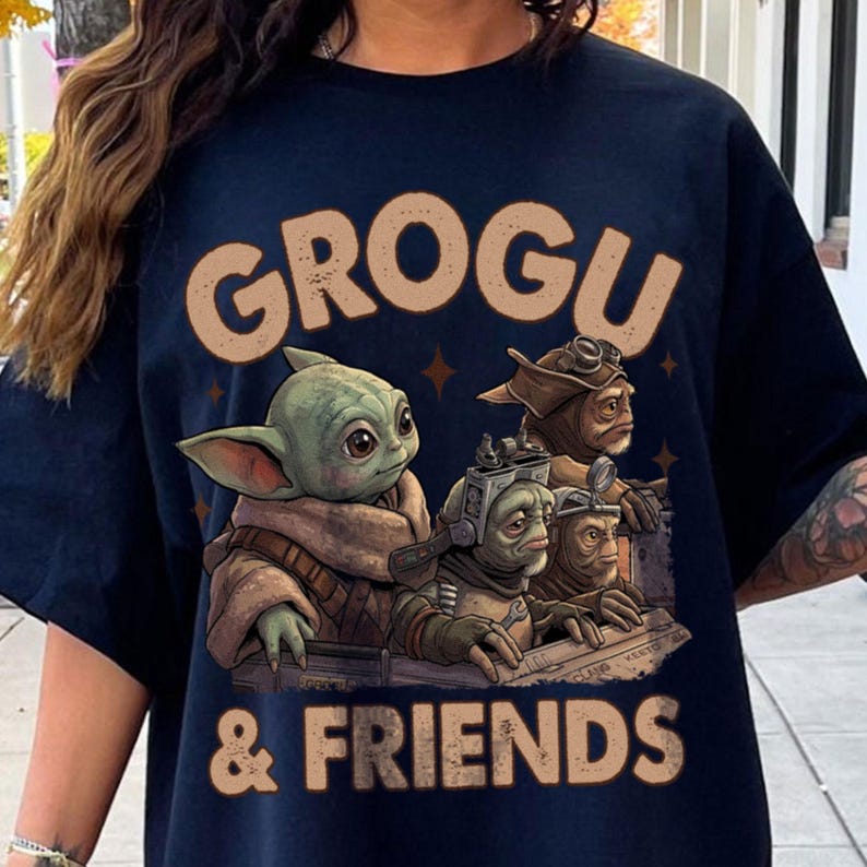 K&ouml;nnte beinhalten: Marineblaues T-Shirt mit dem Text "GROGU & FRIENDS" in beigen Buchstaben. Die Grafik zeigt eine Illustration von Grogu und anderen Charakteren. Das Design ist mittig auf der Vorderseite des T-Shirts platziert.