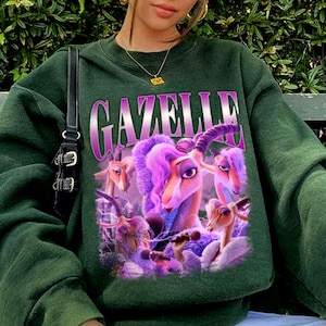 May include: Dark green sweatshirt featuring a graphic of cartoon gazelles in shades of purple and pink. The word "GAZELLE" is printed across the top in a gradient purple font. The sweatshirt has a crew neck and long sleeves.