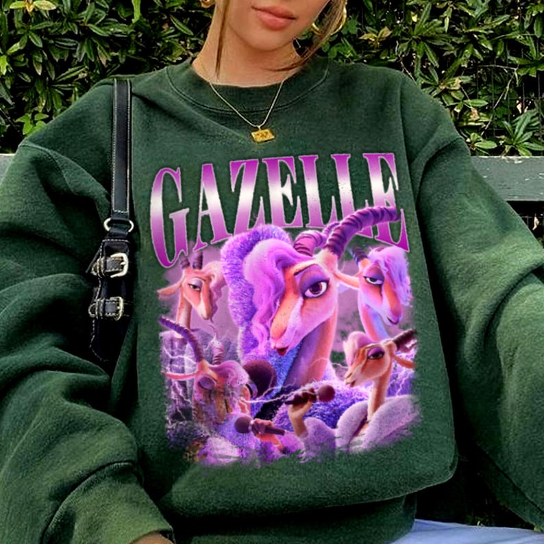 May include: Dark green sweatshirt featuring a graphic of cartoon gazelles in shades of purple and pink. The word "GAZELLE" is printed across the top in a gradient purple font. The sweatshirt has a crew neck and long sleeves.