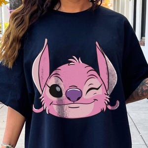 May include: Navy blue t-shirt featuring a pink cartoon character with a winking eye and a smiling expression. The character has large ears and a purple nose. The design is centered on the front of the shirt.