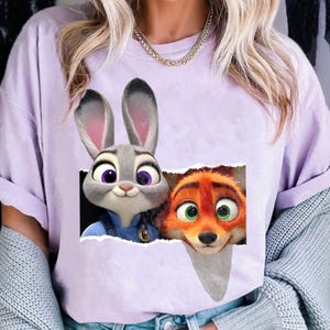 May include: A light purple t-shirt featuring a graphic of Judy Hopps the bunny and Nick Wilde the fox from the animated movie Zootopia. The characters are depicted in a torn paper effect. The shirt is a casual style.