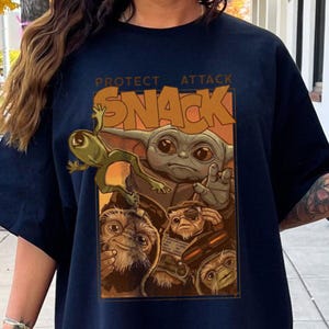 May include: Navy blue t-shirt featuring a graphic of Grogu, a frog-like creature, and other characters from a popular science fiction series. The text "PROTECT ATTACK SNACK" is printed above the characters in a tan color.