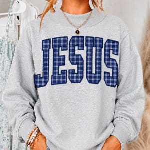 May include: A light gray crewneck sweatshirt featuring the word JESUS in large, block-style letters with a dark blue and white plaid pattern. The garment has long sleeves and a ribbed neckline, paired with a simple gold-toned chain necklace.