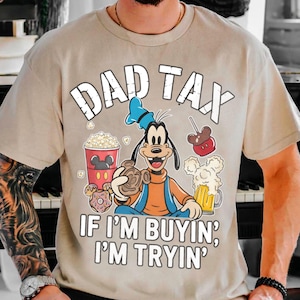 Op de afbeelding: Beige T-shirt met de tekst "DAD TAX" boven een stripfiguur die snacks en drankjes vasthoudt. De zin "IF I'M BUYIN', I'M TRYIN'" staat eronder gedrukt. Het ontwerp bevat popcorn, een Mickey Mouse-vormige appel en een biertje.