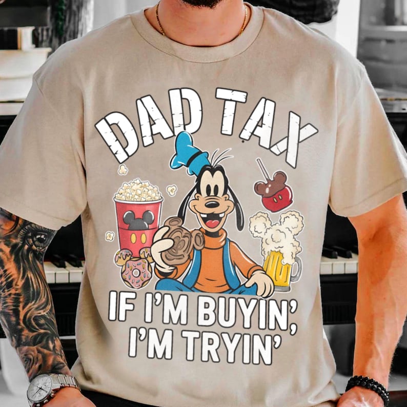 Op de afbeelding: Beige T-shirt met de tekst "DAD TAX" boven een stripfiguur die snacks en drankjes vasthoudt. De zin "IF I'M BUYIN', I'M TRYIN'" staat eronder gedrukt. Het ontwerp bevat popcorn, een Mickey Mouse-vormige appel en een biertje.