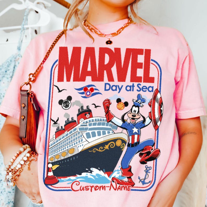 May include: Pink t-shirt featuring a graphic of a cruise ship with the text "MARVEL Day at Sea." The design includes a cartoon character dressed as Captain America. The shirt has a red, white, and blue color scheme.