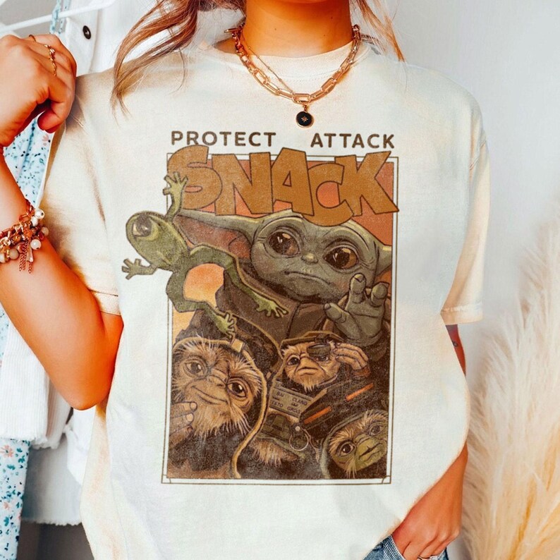 May include: Cream-colored t-shirt featuring a graphic of a character from a popular science fiction franchise. The graphic includes the text "PROTECT ATTACK SNACK" above an illustration of a green frog, a baby-like character, and three other creatures.