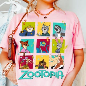 Puede incluir: Camiseta rosa claro con una cuadr&iacute;cula de personajes de Zootopia en cuadrados coloridos. La palabra "Zootopia" est&aacute; impresa en letras verdes en la parte inferior. La camiseta es de manga corta.