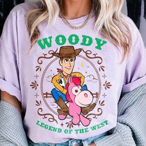 May include: A light purple t-shirt featuring a graphic of the character Woody from Toy Story riding a pink horse. The design includes a brown lasso border, green text that reads WOODY at the top and LEGEND OF THE WEST at the bottom.