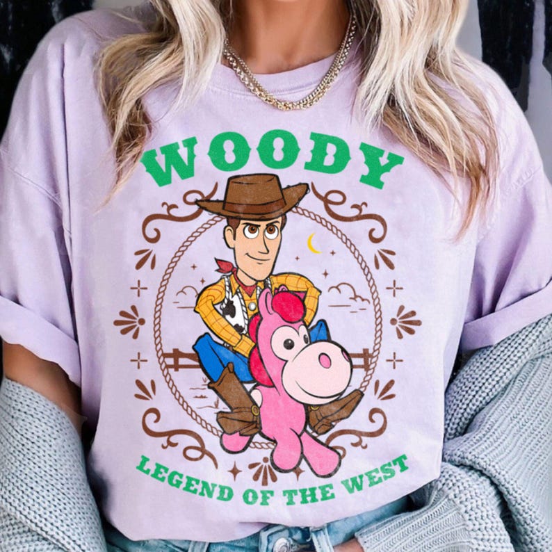 May include: A light purple t-shirt featuring a graphic of the character Woody from Toy Story riding a pink horse. The design includes a brown lasso border, green text that reads WOODY at the top and LEGEND OF THE WEST at the bottom.
