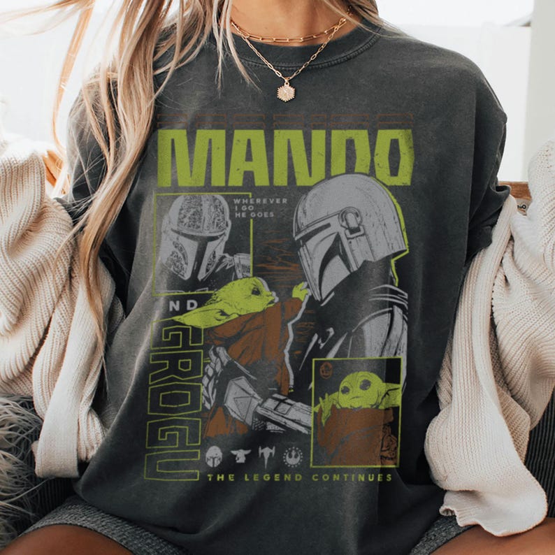 May include: Dark grey sweatshirt with a neon green "MANDO" graphic. The design features illustrations of a Mandalorian helmet and Grogu, with the text "The Legend Continues". The sweatshirt is a casual wear item.