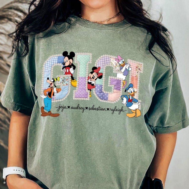 May include: Sage green t-shirt with the word "GIGI" in a patchwork design, featuring Disney characters Mickey Mouse, Minnie Mouse, Goofy, Daisy Duck, and Donald Duck. Names are printed below.