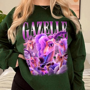 May include: Dark green sweatshirt featuring the word "GAZELLE" in a purple, stylized font. The graphic includes a cartoon image of a gazelle with purple hair and several other gazelles. The sweatshirt has long sleeves and a crew neck.