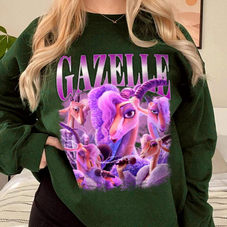 May include: Dark green sweatshirt featuring the word "GAZELLE" in a purple, stylized font. The graphic includes a cartoon image of a gazelle with purple hair and several other gazelles. The sweatshirt has long sleeves and a crew neck.