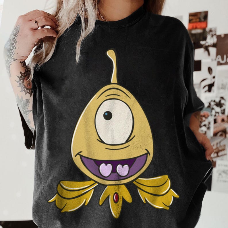 May include: Black t-shirt featuring a yellow cartoon alien with one eye, a purple mouth, and heart-shaped teeth. The alien has yellow wings and a red gem. The shirt is a casual style.
