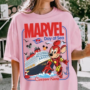 May include: Pink t-shirt featuring a graphic of a cruise ship with a cartoon character in a red and yellow suit. The shirt has the words "MARVEL Day at Sea" in red and blue, with the words "Custom Name" below the ship.