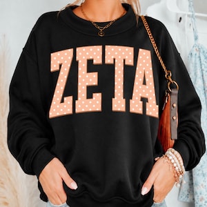 May include: A black long-sleeve sweatshirt featuring the word ZETA in large, block-style letters. The letters are a light peach color with a white polka dot pattern. The sweatshirt has a crew neckline and is worn with a gold chain necklace.