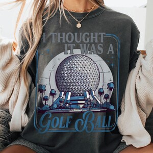 May include: Dark gray sweatshirt with a graphic design featuring a geometric sphere, trees, and the text "I Thought It Was A Golf Ball" in blue and white. The design is framed in a blue rectangle.