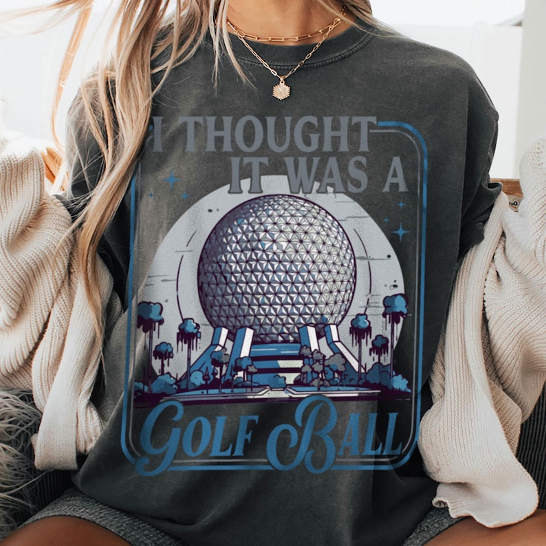 May include: Dark gray sweatshirt with a graphic design featuring a geometric sphere, trees, and the text "I Thought It Was A Golf Ball" in blue and white. The design is framed in a blue rectangle.